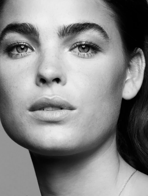 Black and white close-up portrait of a serene adult woman with dark hair, striking eyebrows, and a delicate silver chain necklace.
