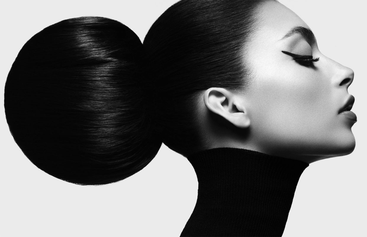 Striking black and white beauty shot of a woman in profile with sleek, dark hair styled in a large bun and distinctive eyeliner.