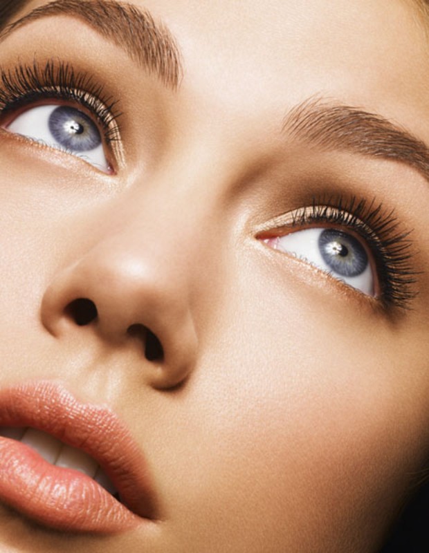 Close-up beauty shot of a woman's face, featuring her eyes with long lashes, eyebrows, nose, and lips, all highlighted with soft, diffused lighting.