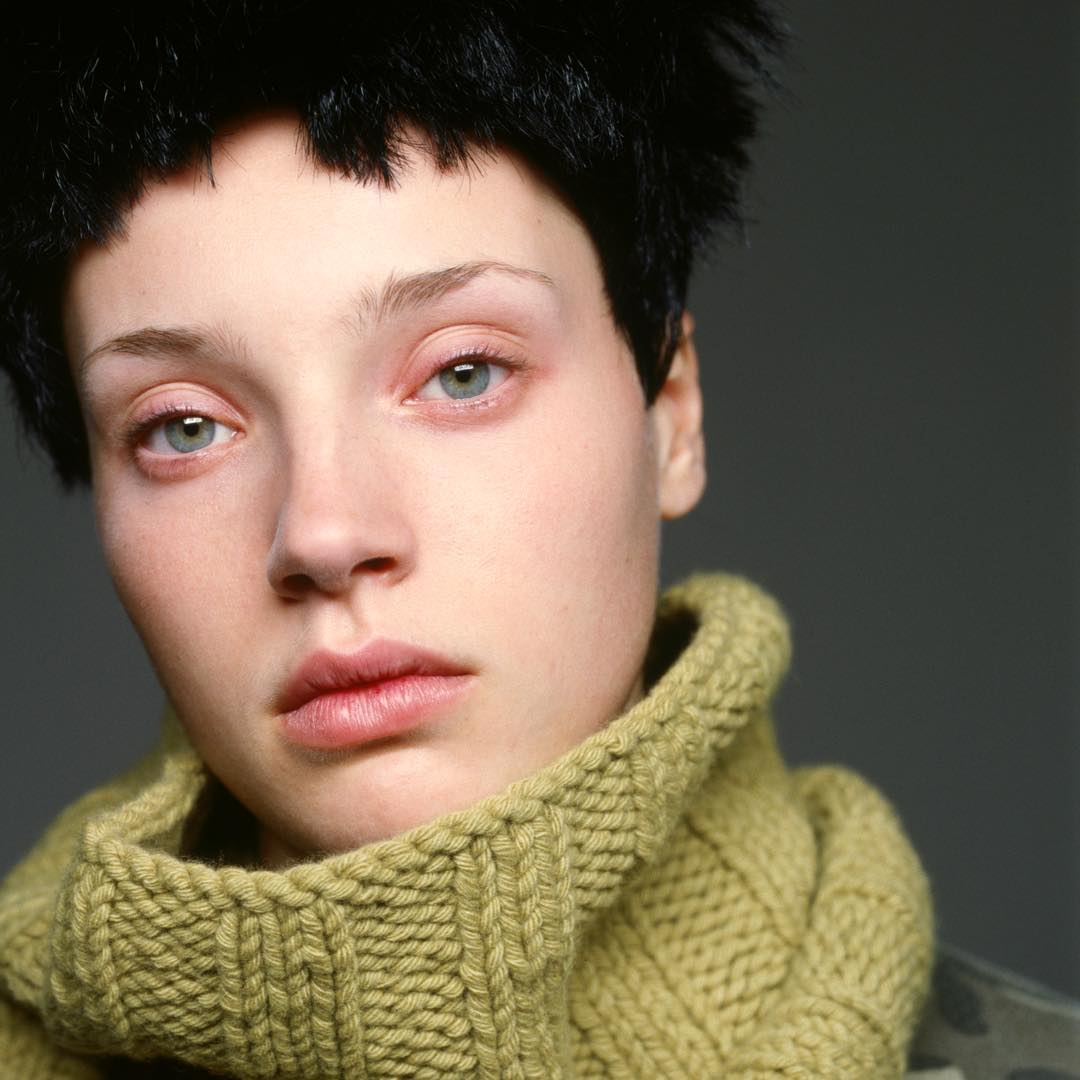 Portrait of a fair-skinned adult female with short black hair wearing a light green knit turtleneck sweater against a grey background.