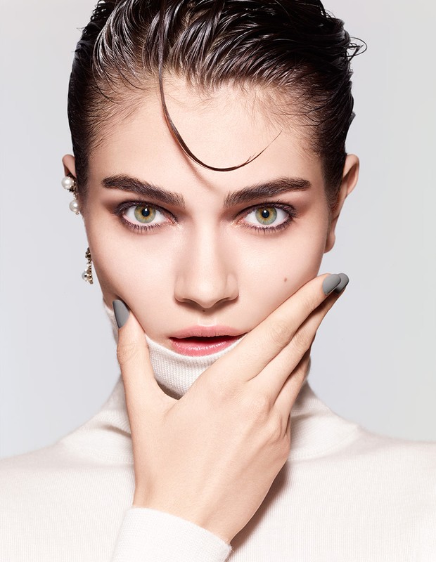 Close-up portrait of a young woman with slicked-back dark hair, green eyes, wearing a white turtleneck, accessorized with pearl earrings.