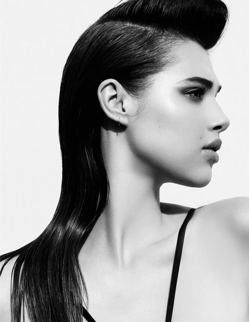 Striking monochrome portrait of a young woman with sleek, dark hair styled in a pompadour, looking to the side against a bright white background.