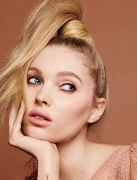 Close-up portrait of a blonde woman with blue eyes, high ponytail, and pink lips resting chin on hand against a warm brown background.
