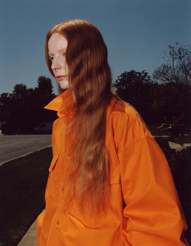 Portrait of a young woman with long red hair wearing an orange button-down shirt, standing outdoors with trees and blue sky.