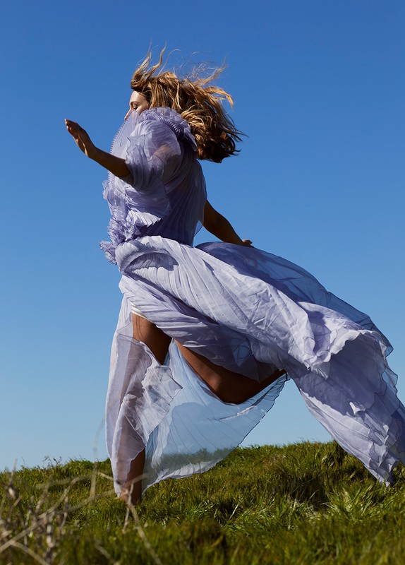 Woman in flowing purple dress leaps across a grassy hill under a clear blue sky, hair blowing in the wind, capturing a sense of freedom.
