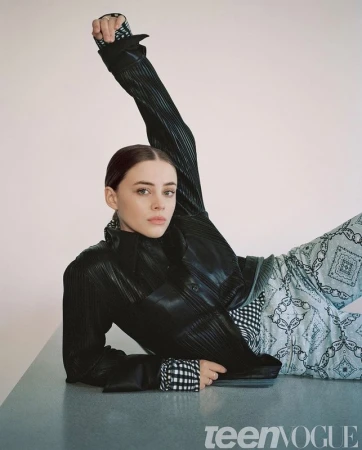 Portrait of a woman reclining on a grey surface wearing a black leather jacket and patterned light blue trousers