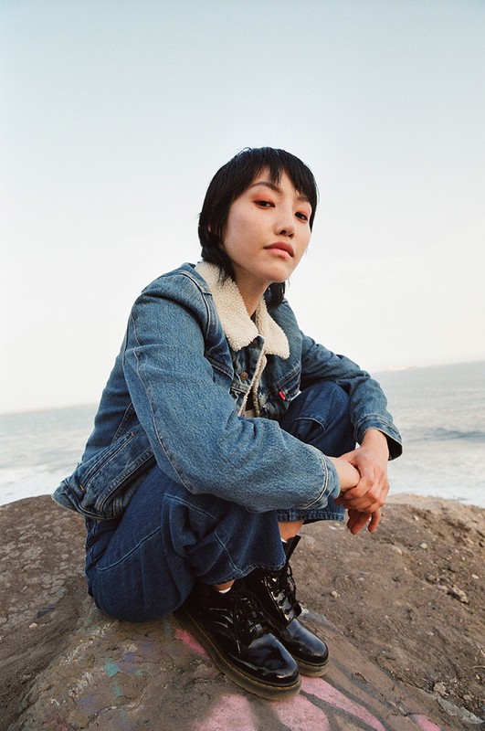 A young woman with short black hair poses outdoors wearing a blue denim jacket, jeans, and black boots, near the ocean and clear sky.