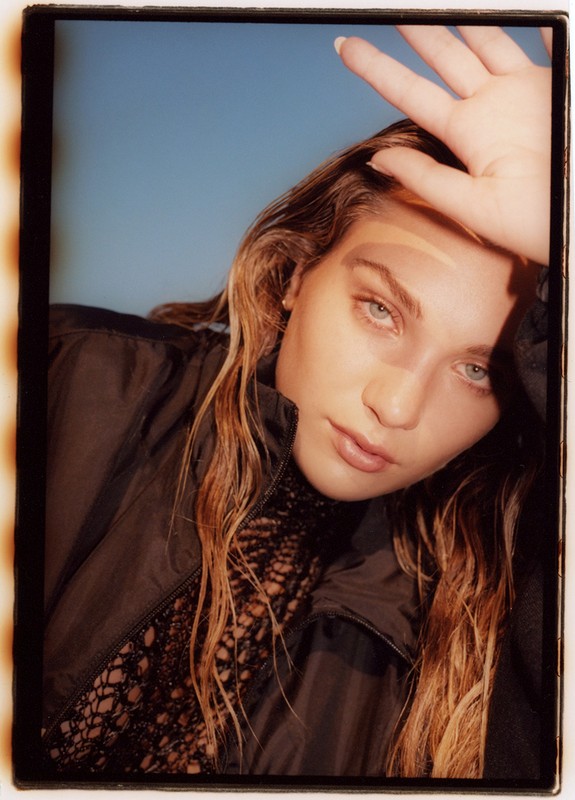 Portrait of a young adult woman with long brown hair shielding her face with her hand, wearing a jacket in front of a blue background.