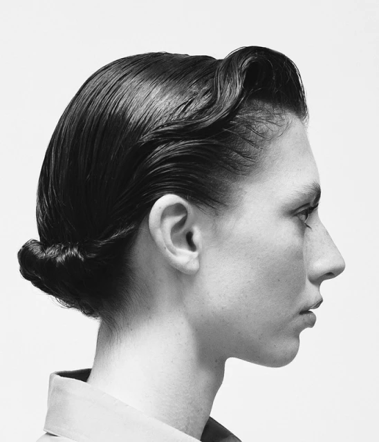 Monochrome studio portrait of a person in profile with slicked-back dark hair styled in a roll and twist, against a white backdrop.