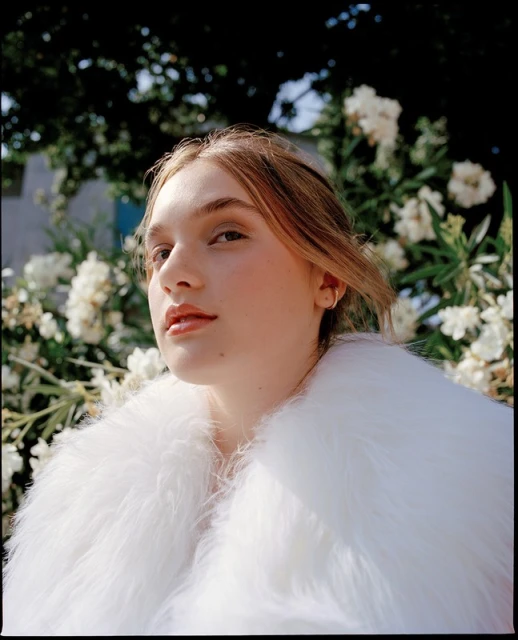 Portrait of a young woman with fair skin and blonde hair wearing a white fur coat, posing in front of white flowers and green foliage.