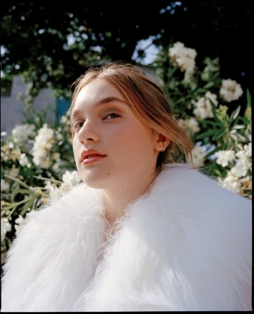 Portrait of a young woman with fair skin and blonde hair wearing a white fur coat, posing in front of white flowers and green foliage.