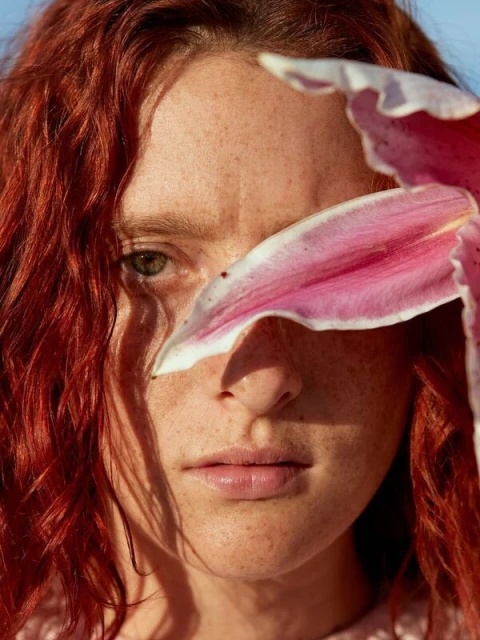 Close-up portrait of an adult woman with red hair, freckled skin, and light eyes, with pink lily petals partially obscuring her face.