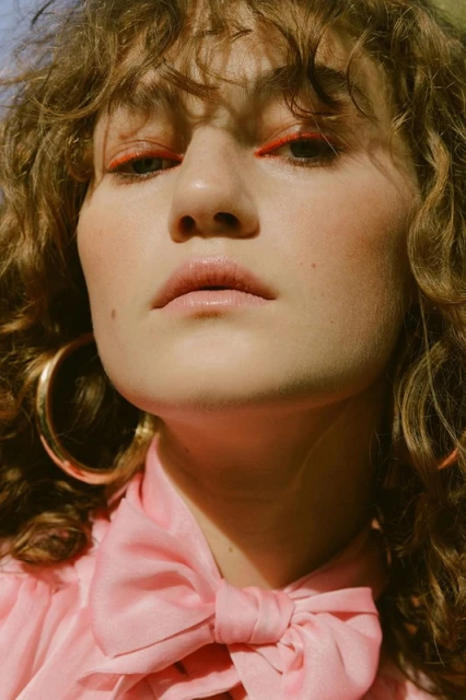 Close-up shot of a woman with curly hair, bright orange eyeliner, a pink bow blouse, and gold hoop earrings in an outdoor setting.