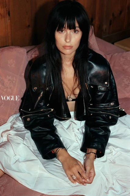 An adult female with long dark hair wearing a black leather jacket sits on a pink sofa in front of a white sheet in a studio setting.