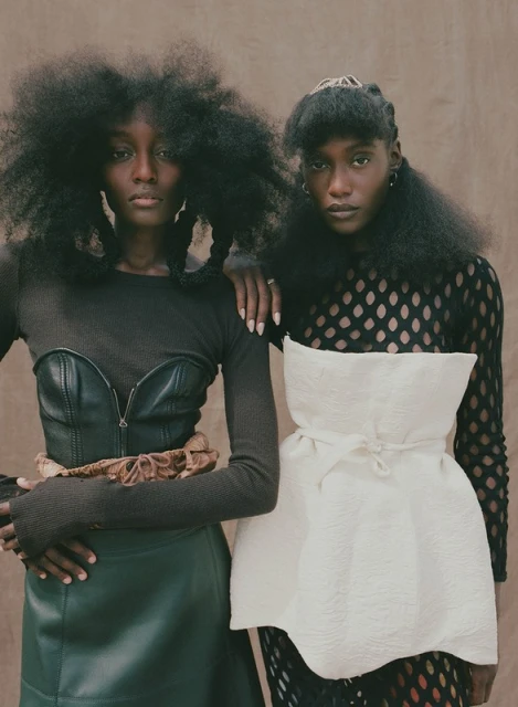 Two Black female models with afros pose indoors. One wears a leather bustier and skirt, the other a white top and sheer black tights.