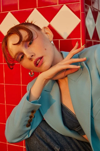 Indoor shot of a woman with reddish-brown hair wearing a blue blazer, jeans and hoop earrings, leaning against a red and white tiled wall.