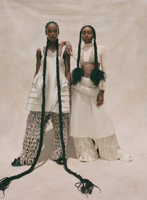 Two models pose in unique off-white outfits with long braids against a plain background in a fashion editorial style studio shot.