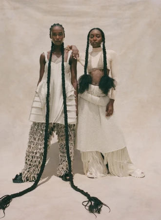 Two Black female models in cream textured outfits with extremely long braided hairstyles standing against a textured backdrop.