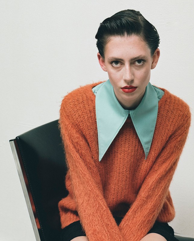Portrait of a fair-skinned woman with dark slicked-back hair, wearing an orange sweater and blue collared shirt, sitting in a black chair.