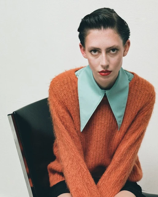 Portrait of a fair-skinned woman with dark slicked-back hair, wearing an orange sweater and blue collared shirt, sitting in a black chair.