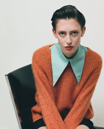 Portrait of a fair-skinned woman with dark slicked-back hair, wearing an orange sweater and blue collared shirt, sitting in a black chair.