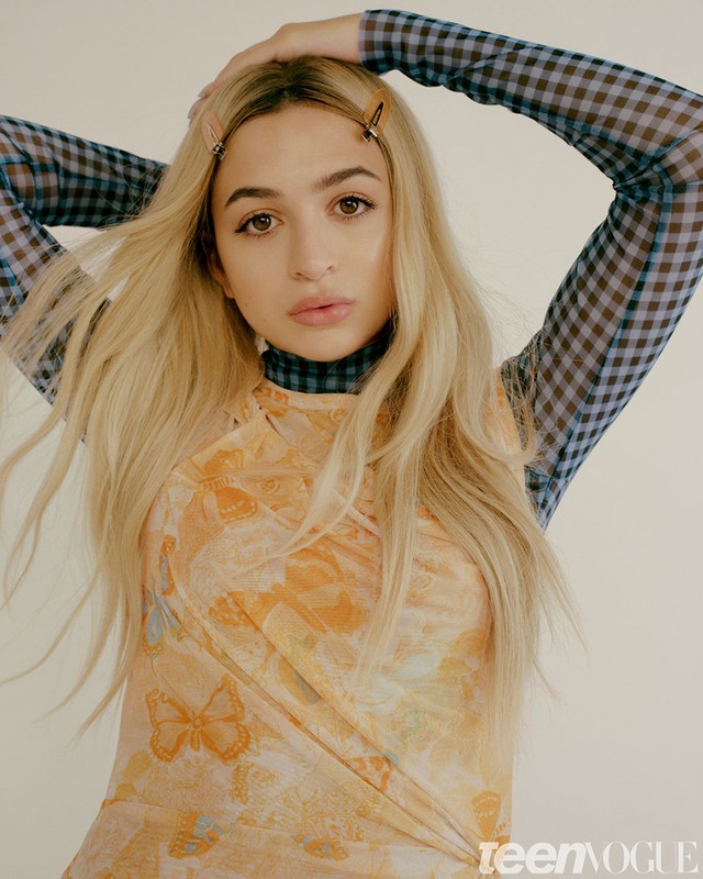 Portrait of a young woman with blonde hair wearing a patterned shirt and dress, hair clips, arms raised slightly, neutral expression.