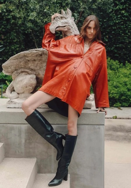 A woman in a red leather jacket and black boots poses on stone steps next to a stone griffin statue in an outdoor garden setting.