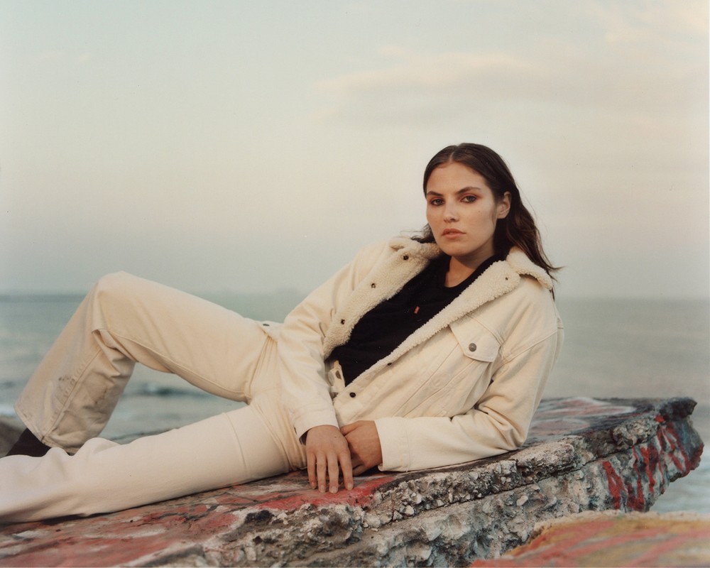 A young woman with brown hair wearing a jacket and jeans, lying on a concrete ledge in front of a body of water under an overcast sky.