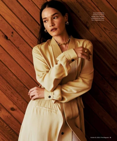 Woman in pale yellow trench coat leaning against diagonal wooden slat wall wearing chunky gold necklace