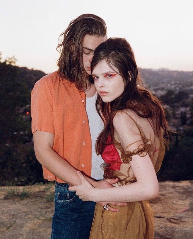 A young couple embraces in a sunlit outdoor setting, showcasing an orange button-down shirt, a flowing dress, and a natural background.