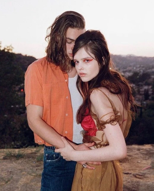 A young couple embraces in a sunlit outdoor setting, showcasing an orange button-down shirt, a flowing dress, and a natural background.