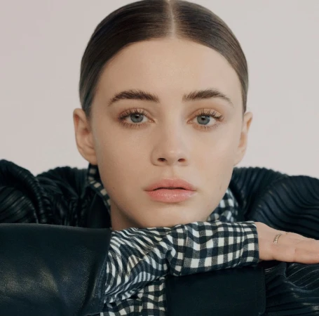 Close-up portrait of a woman resting her chin on her arms, wearing a black leather jacket and a black and white gingham shirt.