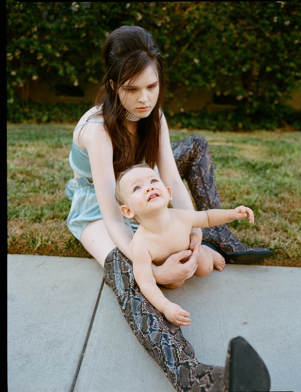 Woman in snake-print boots sits with a baby in her lap, outdoors on a sunny day with green foliage in the background. Fashion photography.