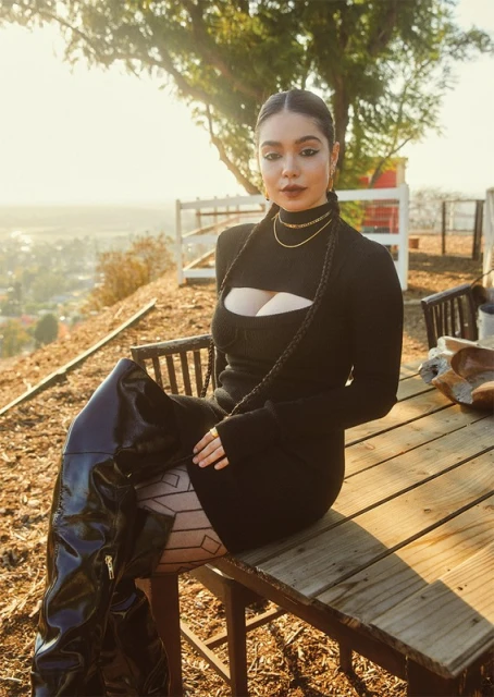 Portrait of a woman in a black dress and shiny black thigh-high boots sitting on a wooden table outside with a city view.