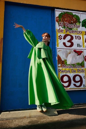 An adult woman wearing a vibrant green dress stands in an outdoor setting with a food advertisement backdrop in this eye-catching fashion photo.