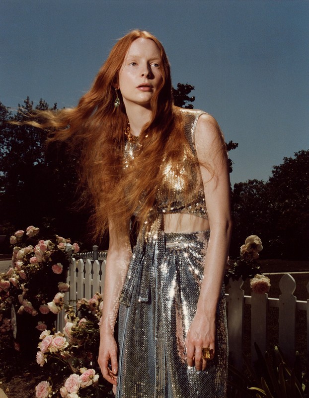 Portrait of a red-haired woman in shimmering gold and silver attire outdoors against a pale blue sky and garden backdrop with a white picket fence.