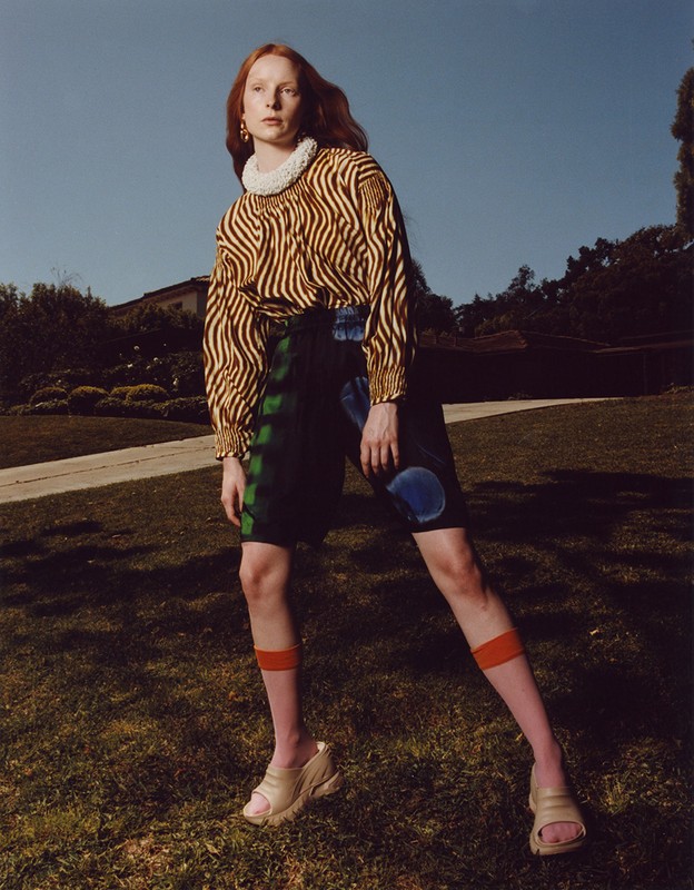 Fashion shot of a red-haired woman in a striped top and patterned shorts, posing outdoors in a grassy area with a clear blue sky.