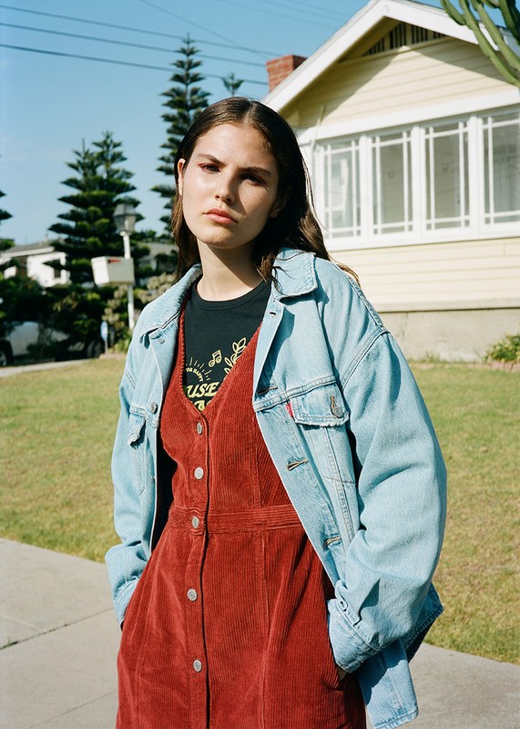 A young woman with auburn hair poses in front of a light yellow house wearing a denim jacket over a rust corduroy dress.