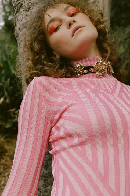 Fashion shot of a young woman with curly hair wearing a patterned pink top and ornamental gold collar leaning against a tree outdoors.