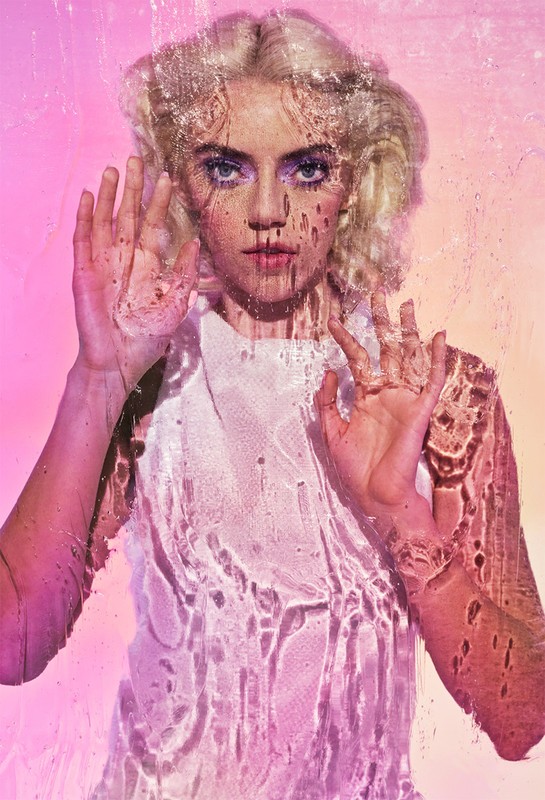 Young woman with blonde hair poses behind textured glass in a close-up shot with pink-to-yellow gradient background in a beauty context.