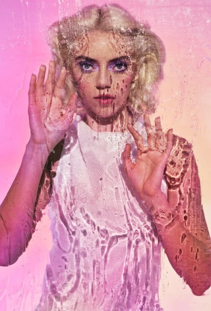 Young woman with blonde hair poses behind textured glass in a close-up shot with pink-to-yellow gradient background in a beauty context.