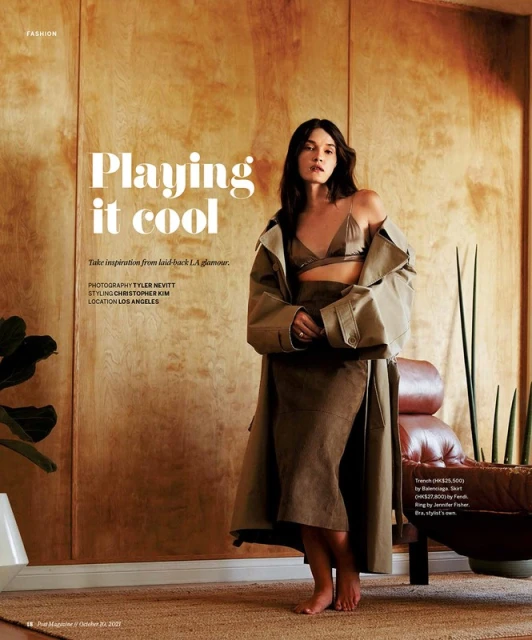Fashion editorial shot of a slender woman in a brown bra and skirt, wearing a long coat, posing in front of a wood paneled wall.