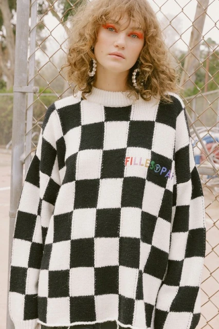 An adult female with curly hair and orange eyeshadow poses in front of a chainlink fence while wearing a checkerboard sweater.