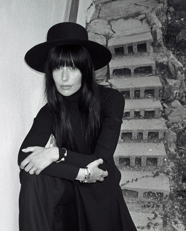 Monochrome portrait of a woman wearing a wide-brimmed black hat and turtleneck, seated on stone steps outdoors.