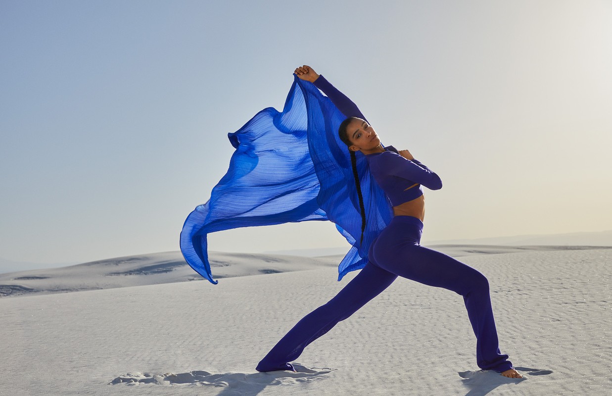 A woman in athletic wear poses with a flowing blue scarf in a yoga-like stance in a sandy desert landscape with a clear sky backdrop.