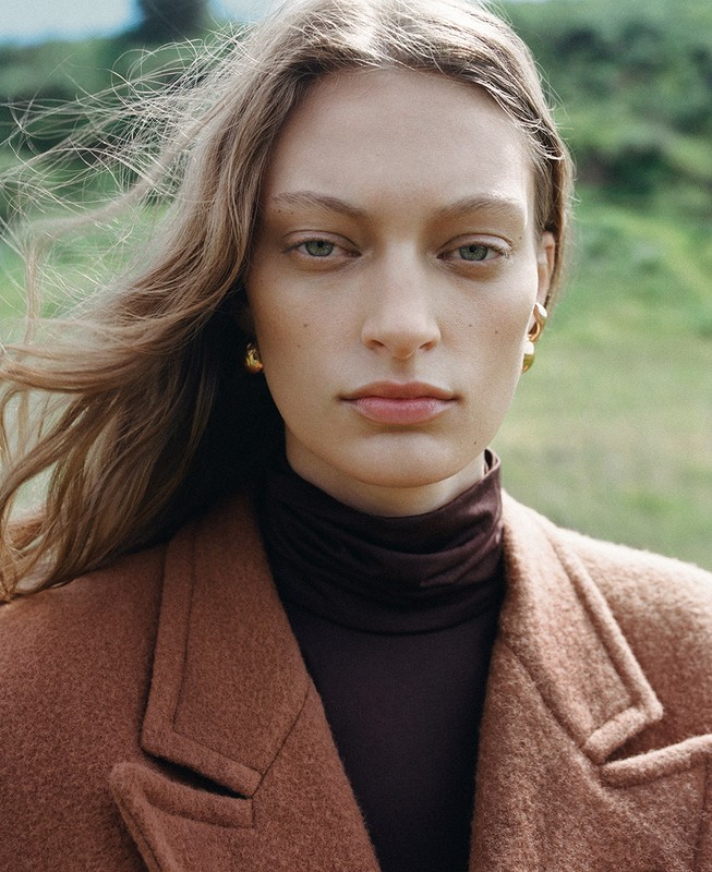 Close-up portrait of a woman with long brown hair, wearing a brown wool coat and gold earrings, against an outdoor green background.