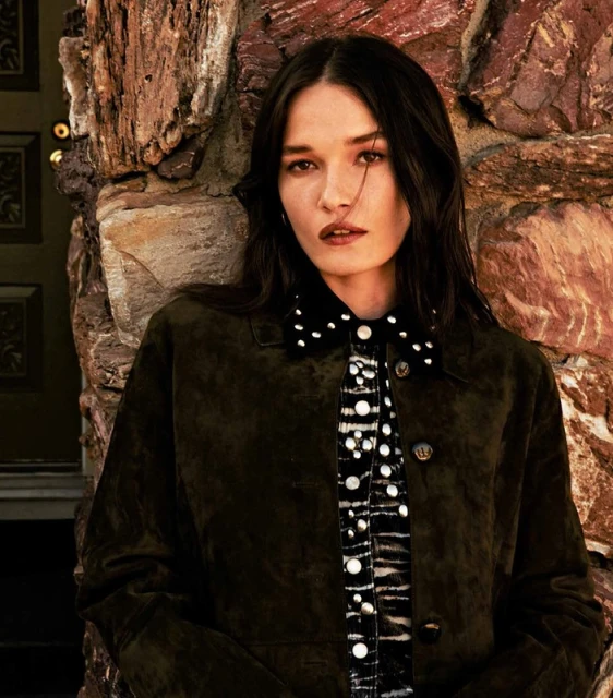 Portrait of a woman with dark hair in a patterned shirt and jacket, leaning against a weathered stone wall indoors.