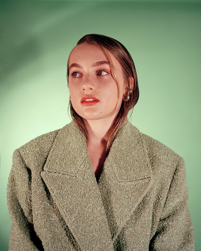 Portrait of a young woman with slicked-back hair wearing a textured green jacket, looking off to the side against a green background.