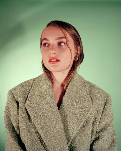 Portrait of a young woman with slicked-back hair wearing a textured green jacket, looking off to the side against a green background.
