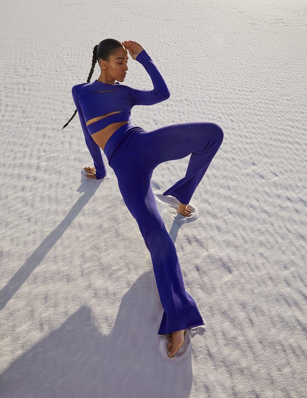 Woman in blue knitwear poses in a white desert landscape. She wears a long-sleeved crop top and flared pants, hand raised to head.